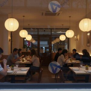 Вечерний вид на зал ресторана из‑за окна, видны накрытые столы и гости. Вечерний вид на зал ресторана из‑за окна